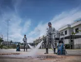 Uma pessoa usando equipamento de segurança usa uma lavadora de alta pressão para limpar o pavimento ao ar livre.