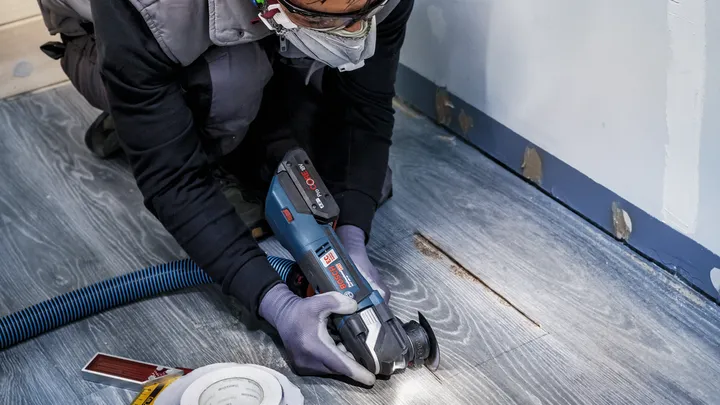 Pessoa usando equipamento de segurança corta um piso de madeira com uma ferramenta elétrica.