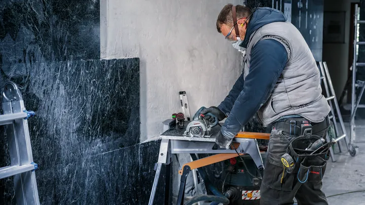 Uma pessoa usando equipamento de segurança corta ladrilhos de pedra usando uma ferramenta elétrica em uma bancada de trabalho.