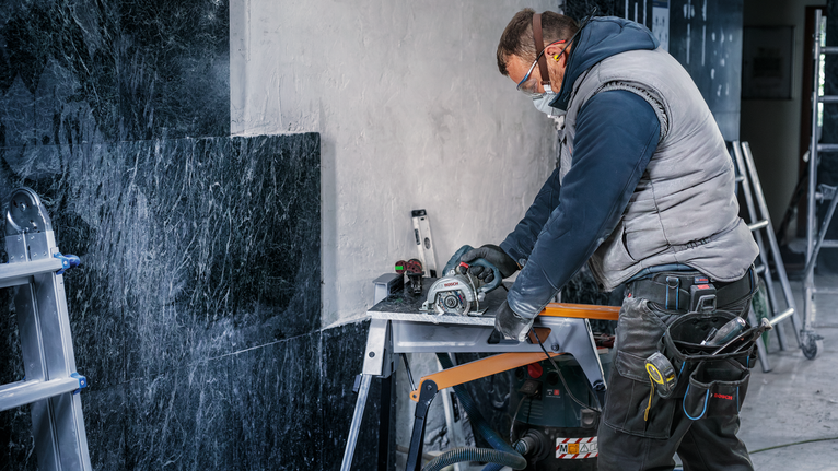 Uma pessoa usando equipamento de segurança corta ladrilhos de pedra usando uma ferramenta elétrica em uma bancada de trabalho.