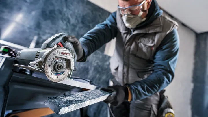 Uma pessoa usando equipamento de segurança corta um azulejo com uma ferramenta elétrica em um ambiente interno.