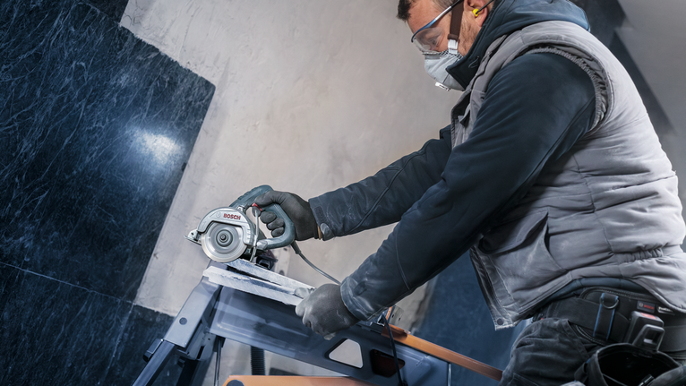 Uma pessoa usando equipamento de segurança corta azulejos com uma serra circular em uma bancada de trabalho.
