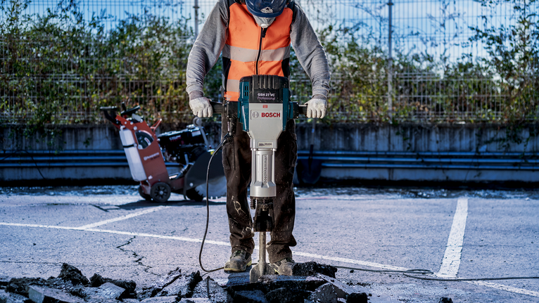 Uma pessoa usando equipamento de segurança usa um martelo demolidor para quebrar pavimento de concreto.