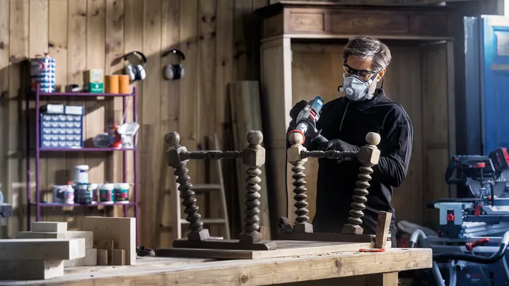 Uma pessoa usando equipamento de segurança fura uma mesa de madeira virada de cabeça para baixo em uma oficina.