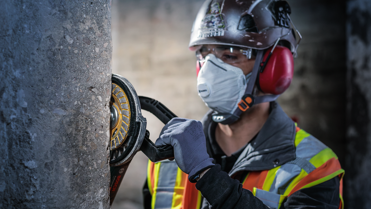 Uma pessoa usando equipamento de segurança tritura um pilar de concreto com uma ferramenta elétrica.