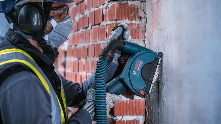 Trabalhador usando equipamento de segurança usa uma ferramenta elétrica para remover argamassa de uma parede de tijolos.