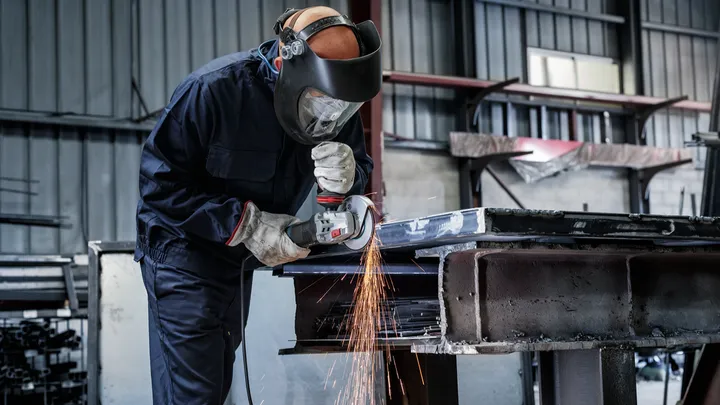 Uma pessoa usando equipamento de segurança tritura vigas de metal, produzindo faíscas brilhantes.