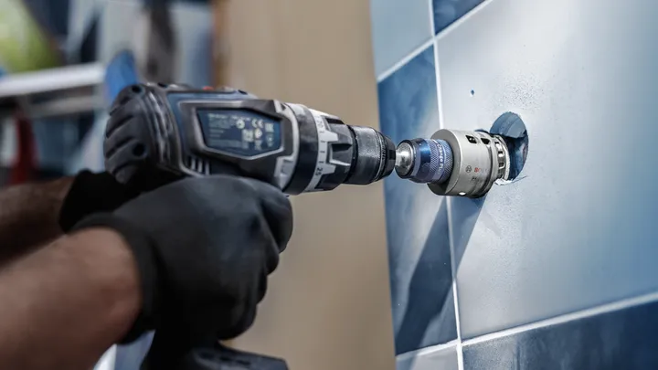 Uma pessoa usando equipamento de segurança faz um furo circular em uma parede de azulejos.