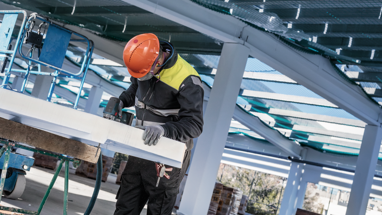 Uma pessoa usando equipamento de segurança perfura um grande painel branco em um canteiro de obras.