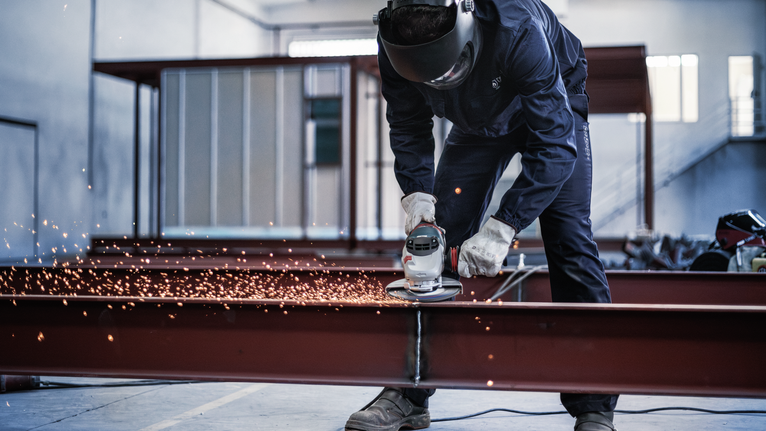 Uma pessoa usando equipamento de segurança tritura uma viga de metal, produzindo faíscas.