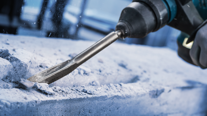 Uma pessoa usando equipamento de segurança usa um martelo rotativo para cinzelar concreto.