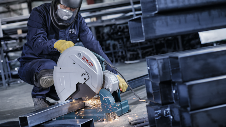 Um trabalhador usando equipamento de segurança corta uma viga de metal com uma serra elétrica, produzindo faíscas.