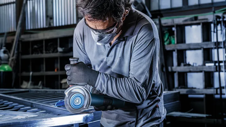 Uma pessoa usando equipamento de segurança tritura uma superfície metálica com uma ferramenta elétrica.