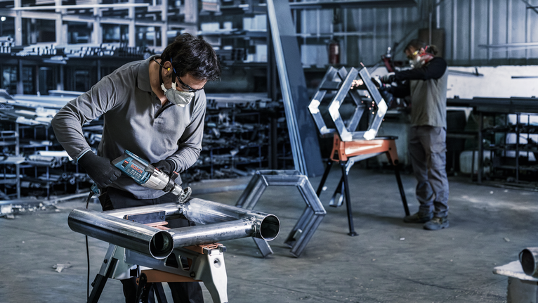 Duas pessoas em uma oficina usando ferramentas elétricas em estruturas de metal, ambas usando equipamentos de segurança.