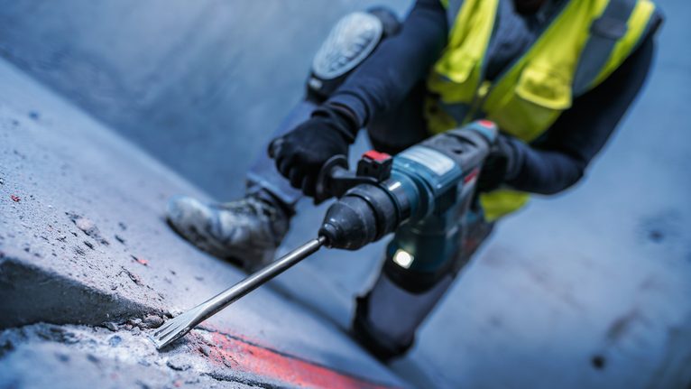 Uma pessoa usando equipamento de segurança usa um martelo rotativo para quebrar concreto.