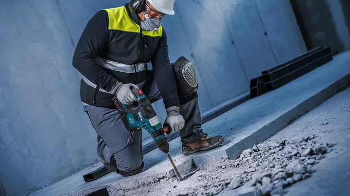Uma pessoa usando equipamento de segurança usa um martelo rotativo para quebrar concreto.