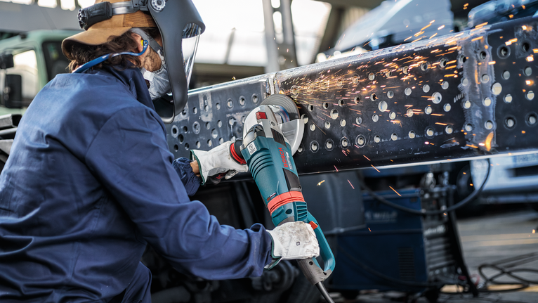 Uma pessoa usando equipamento de segurança tritura uma viga de metal, gerando faíscas brilhantes.