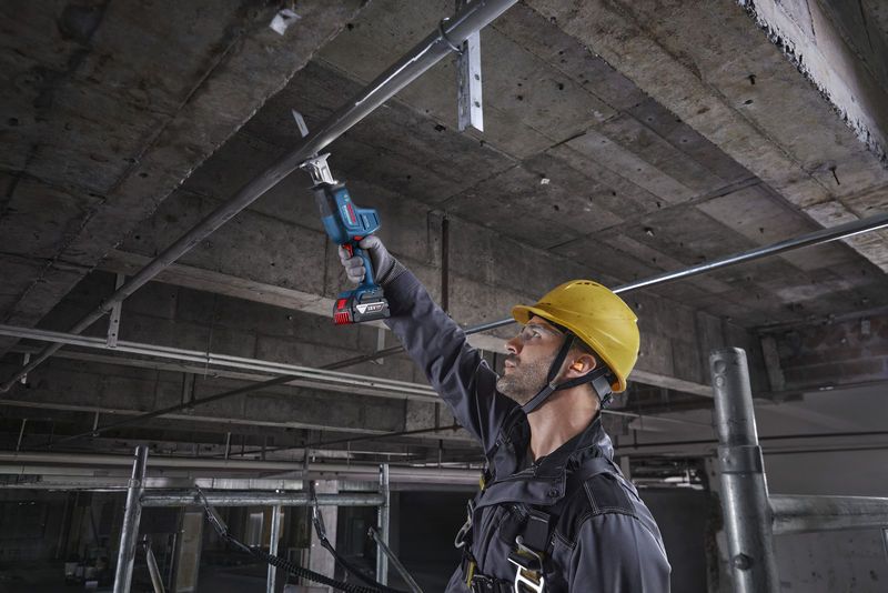 Uma pessoa usando equipamento de segurança usa uma serra alternativa para cortar um cano de metal acima da cabeça.