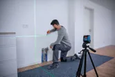 Uma pessoa ajoelhada marcando uma parede guiada por uma ferramenta de nivelamento a laser em um tripé.