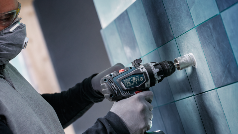 Uma pessoa usando equipamento de segurança perfura uma parede revestida de azulejos.