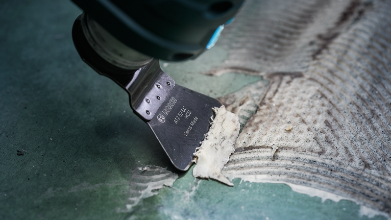 Ferramenta multifuncional oscilante para raspar resíduos de adesivo de um piso texturizado verde.