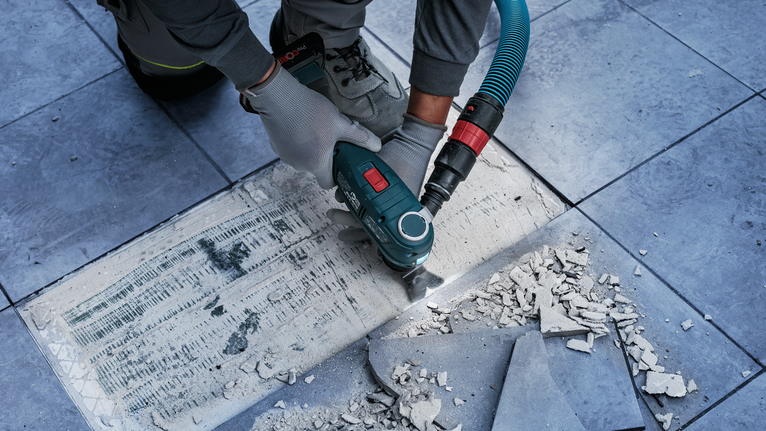 Uma pessoa usando equipamento de segurança remove adesivo para azulejos com uma ferramenta elétrica em um piso revestido com azulejos.