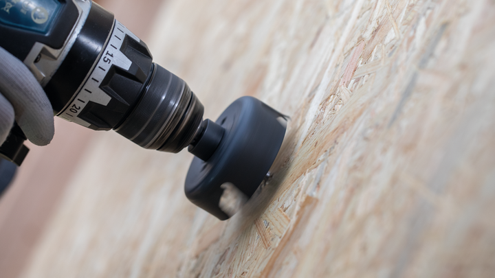 Pessoa usando equipamento de segurança faz um grande furo na madeira.