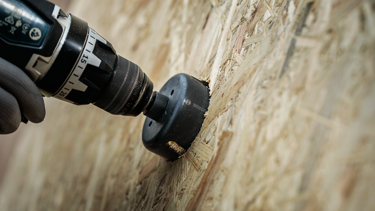 Pessoa usando equipamento de segurança utilizando uma serra copo para perfurar uma placa de OSB (Oriented Strand Board).