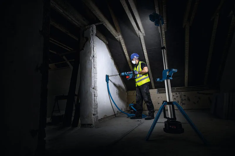 Uma pessoa usando equipamento de segurança perfura uma parede, iluminada por uma luz de obra sem fio.