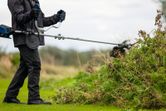 Uma pessoa usando equipamento de segurança apara arbustos grossos com uma roçadeira ao ar livre.
