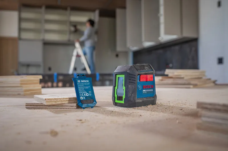 Ferramenta de nivelamento a laser sobre uma mesa empoeirada; pessoa trabalhando em uma escada ao fundo.
