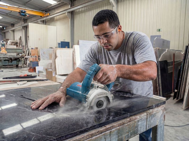 Uma pessoa usando equipamento de segurança utiliza uma cortadora de azulejos diamantada em uma placa de mármore preto.