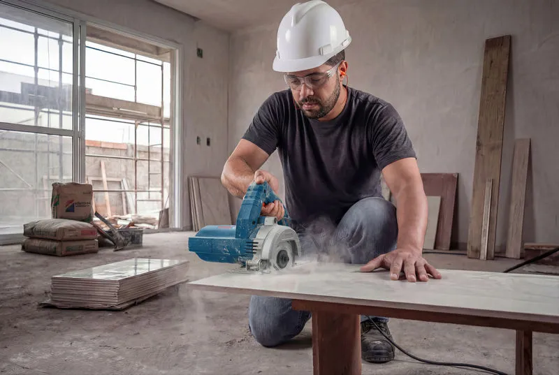 Uma pessoa usando equipamento de segurança utiliza uma cortadora de azulejos diamantada para cortar um azulejo cerâmico.