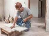 Um trabalhador usando equipamento de segurança utiliza uma cortadora de azulejos diamantada para cortar uma peça de cerâmica.