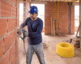 Uma pessoa usando equipamento de segurança utiliza um cortador de azulejos diamantado em uma parede de tijolos.