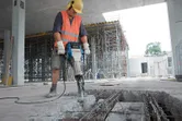 Uma pessoa usando equipamento de segurança usa um martelo demolidor para demolir concreto em um canteiro de obras.