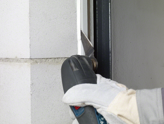 Um trabalhador usando equipamento de segurança corta o selante de uma moldura de janela com uma ferramenta elétrica.