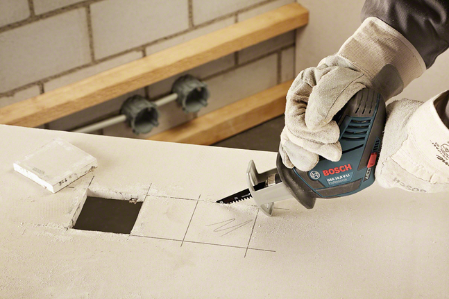 Trabalhador usando equipamento de segurança usa uma serra alternativa para cortar um furo quadrado na parede de gesso.