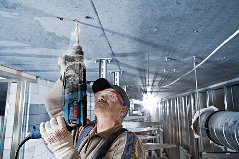 Uma pessoa usando equipamento de segurança perfura um teto de concreto em um ambiente industrial.