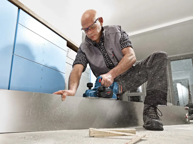 Una persona que usa equipo de seguridad cepilla una tabla de madera utilizando una cepilladora inalámbrica.