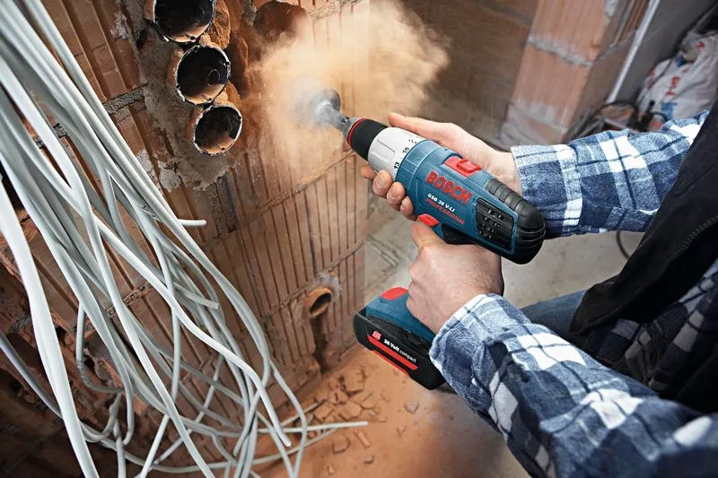 Una persona perfora agujeros en una pared de ladrillos rodeada de cables eléctricos.