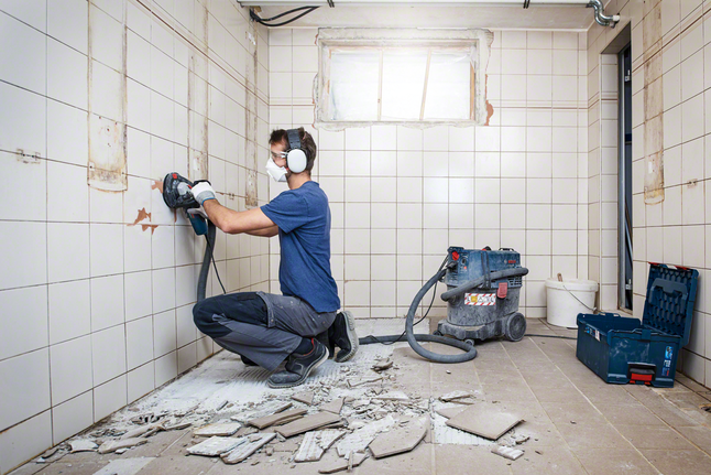 Una persona que usa equipo de seguridad quita el adhesivo para baldosas de una pared revestida de azulejos en una escena de renovación.