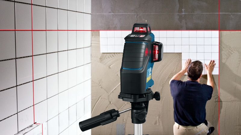 La herramienta de nivelación láser proyecta líneas rojas mientras una persona instala azulejos en una pared.