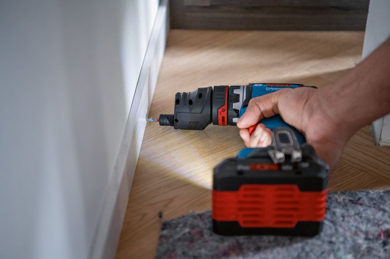 Una persona perfora un zócalo usando un taladro inalámbrico sobre un piso de madera.