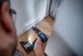 Persona que utiliza un taladro inalámbrico para fijar un tornillo en un piso de madera cerca de una pared.