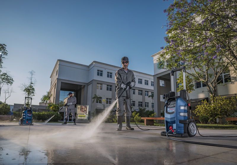 Dos personas que usan equipo de seguridad limpian el pavimento al aire libre utilizando una hidrolavadora.