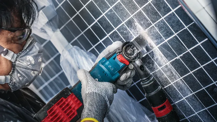 Una persona que usa equipo de seguridad perfora una pared de azulejos generando polvo.
