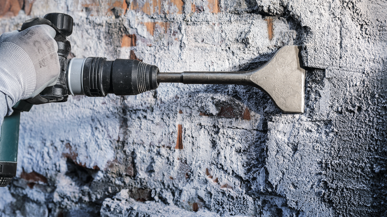 Una persona que usa equipo de seguridad utiliza un taladro cincel para quitar yeso de una pared de ladrillos.