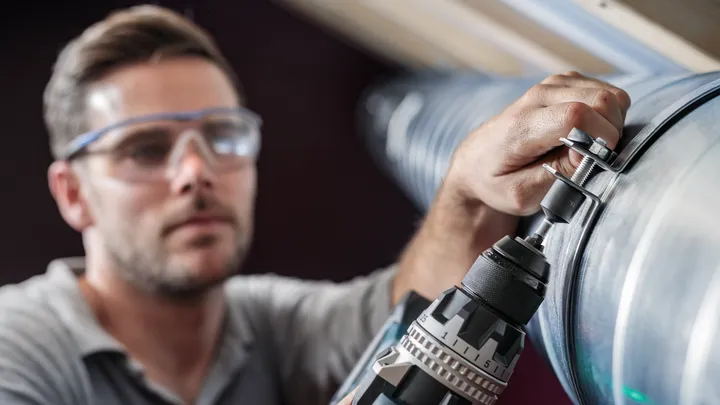 Una persona que usa equipo de seguridad fija un perno a un conducto de metal con un taladro eléctrico.