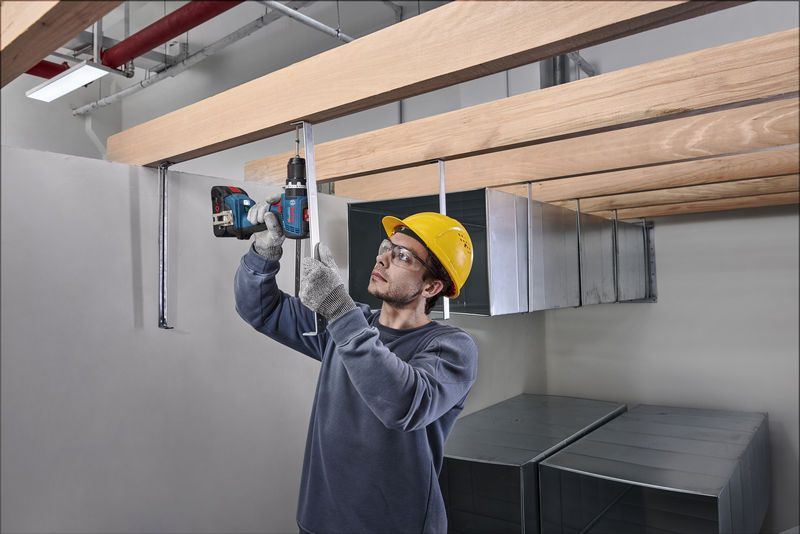 Una persona que usa equipo de seguridad perfora una varilla de metal debajo de vigas de madera.